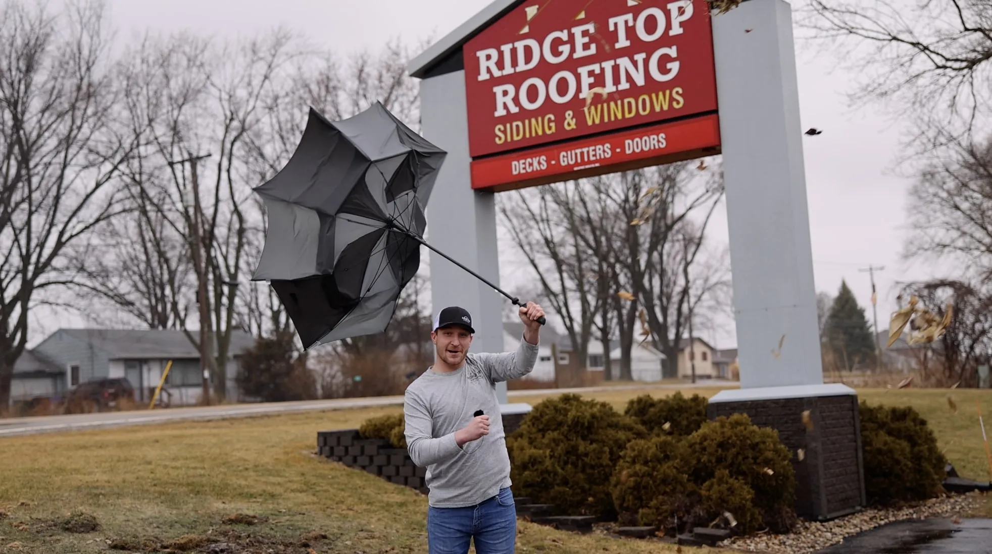 Man holding an inside-out umbrella standing in front of a Ridge Top Roofing sign on a windy day.